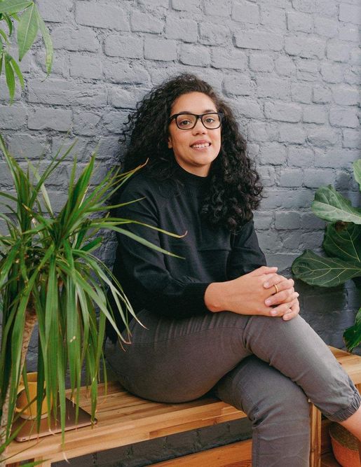Photo of Alex Harris, the CEO and Artistic Director of Beyond Face, looking and smiling directly at the camera while sitting on a wooden bench with her legs crossed and her hands on her knees with a plant either side of her