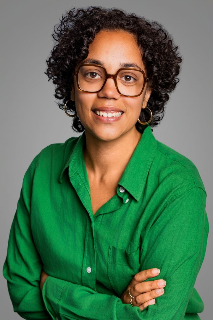 Headshot of a woman with short, curly, brown hair and square glasses. She is wearing a green top, has her arms crossed and smiles at the camera.