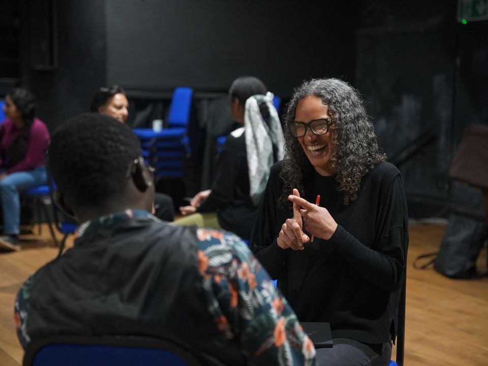 A person with curly gray hair and glasses sits in a chair, smiling and gesturing while engaging in conversation with a man wearing a floral shirt. Others are seated in the background, with the setting appearing to be a rehearsal space.