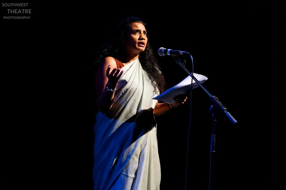 A person in a light-coloured sari passionately delivers a speech into a microphone on a dark stage, holding papers and gesturing expressively.