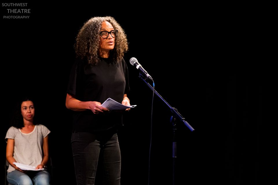Person with curly hair and glasses speaking at a microphone on a dark stage, holding notes, with another seated performer in the background.