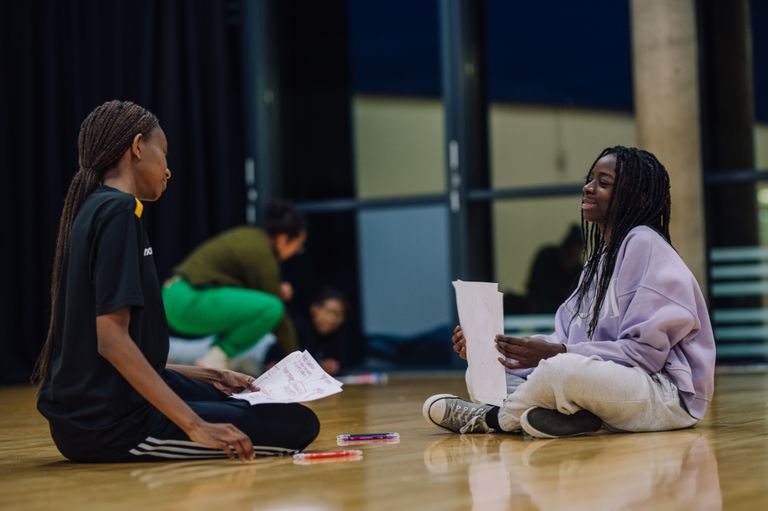 Two young people sit on the floor smiling and holding scripts, there is a adult facilitating in the background speaking with another young person