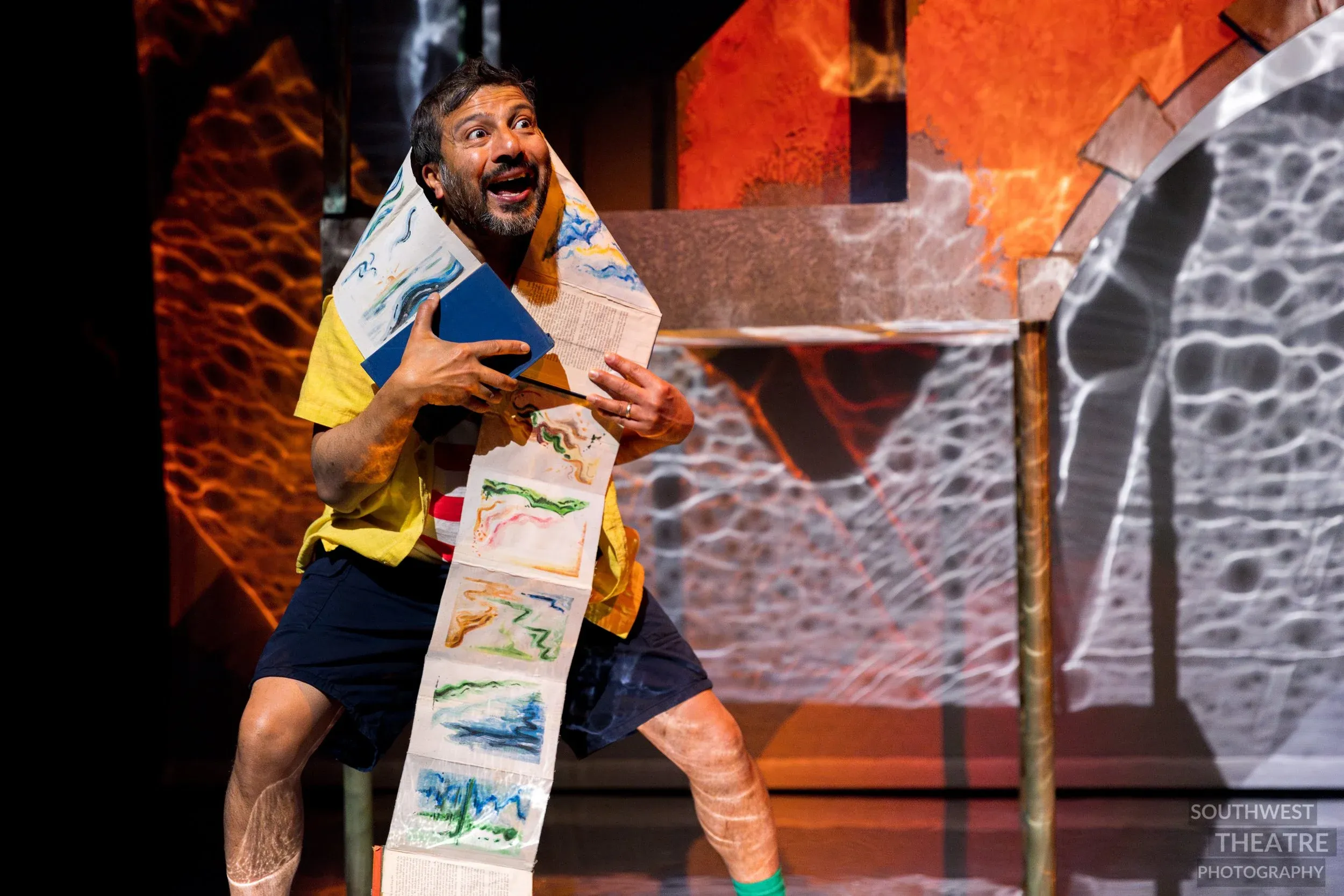 A man on stage is dressed in a red and white striped shirt and is holding books, he was an inquisitive expression.