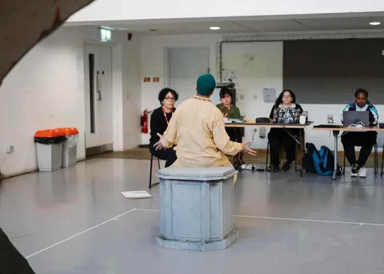 A man dressed in thobe and kufi is in a rehearsal room performing to three woman at a desk