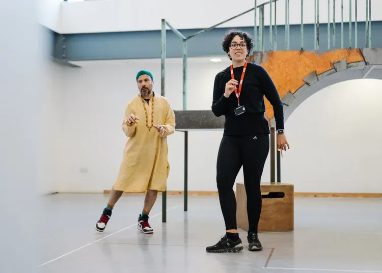 A man dressed in a thobe and kufi dances in a rehearsal room. There is a woman in all black with a lanyard on stood in front of him dancing in the same way