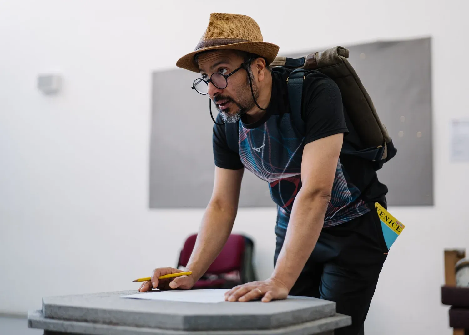 A man is wearing a boater hat, glasses and a backpack he is in a rehearsal studio and is leaning against a table looking at someone out of frame