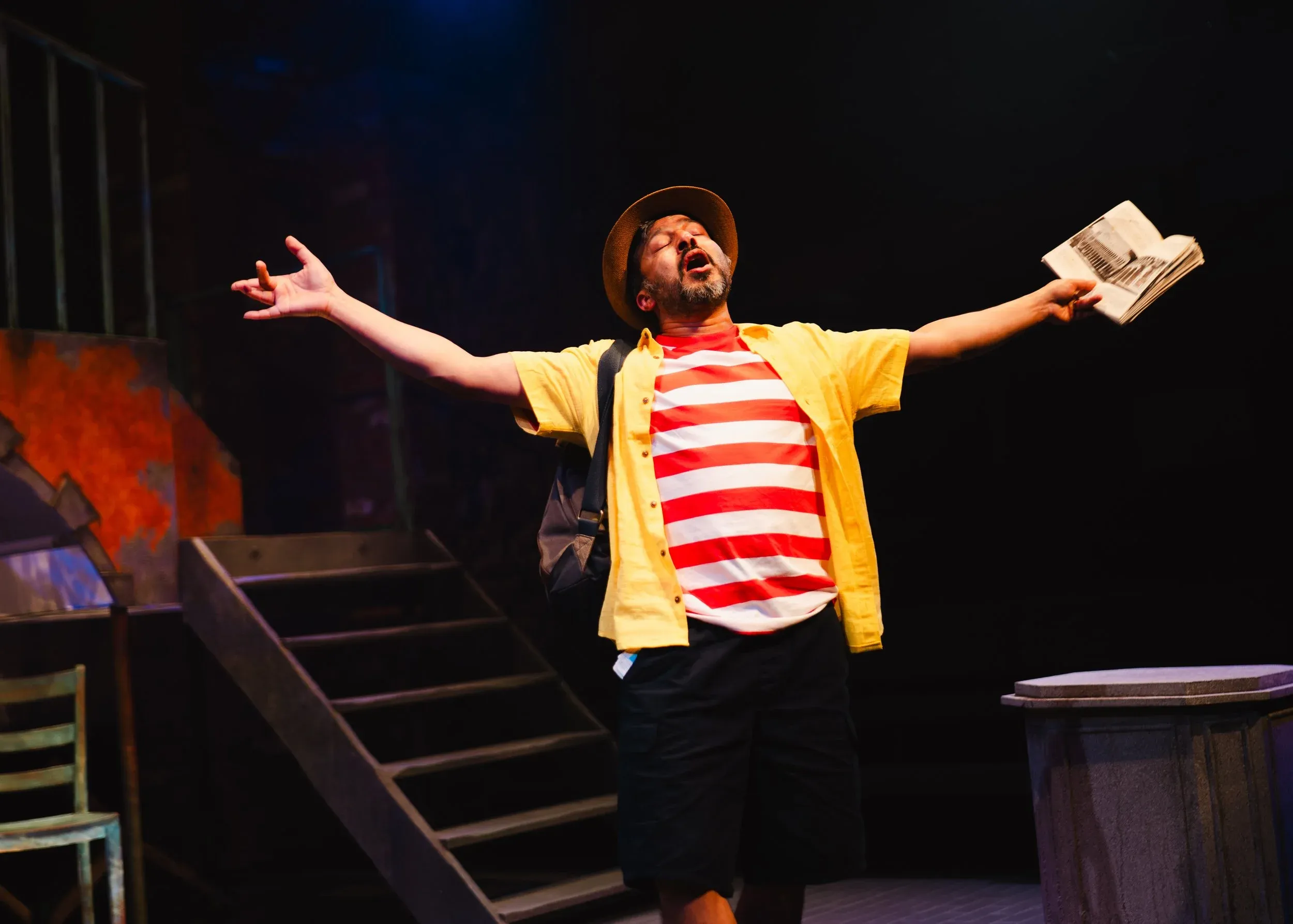 A man on stage is dressed in a red and white striped shirt with a hat on, he is has his arms up to the sky and eyes shut