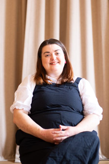 Photo of Helen Bovey, the Operations Manager of Beyond Face, sitting on a seat in front of a curtain and looking directly at the camera and smiling with her hands crossed on her knees
