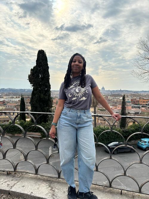 A black woman in braids with denim jeans and a grey t-shirt leans against a fence smiling