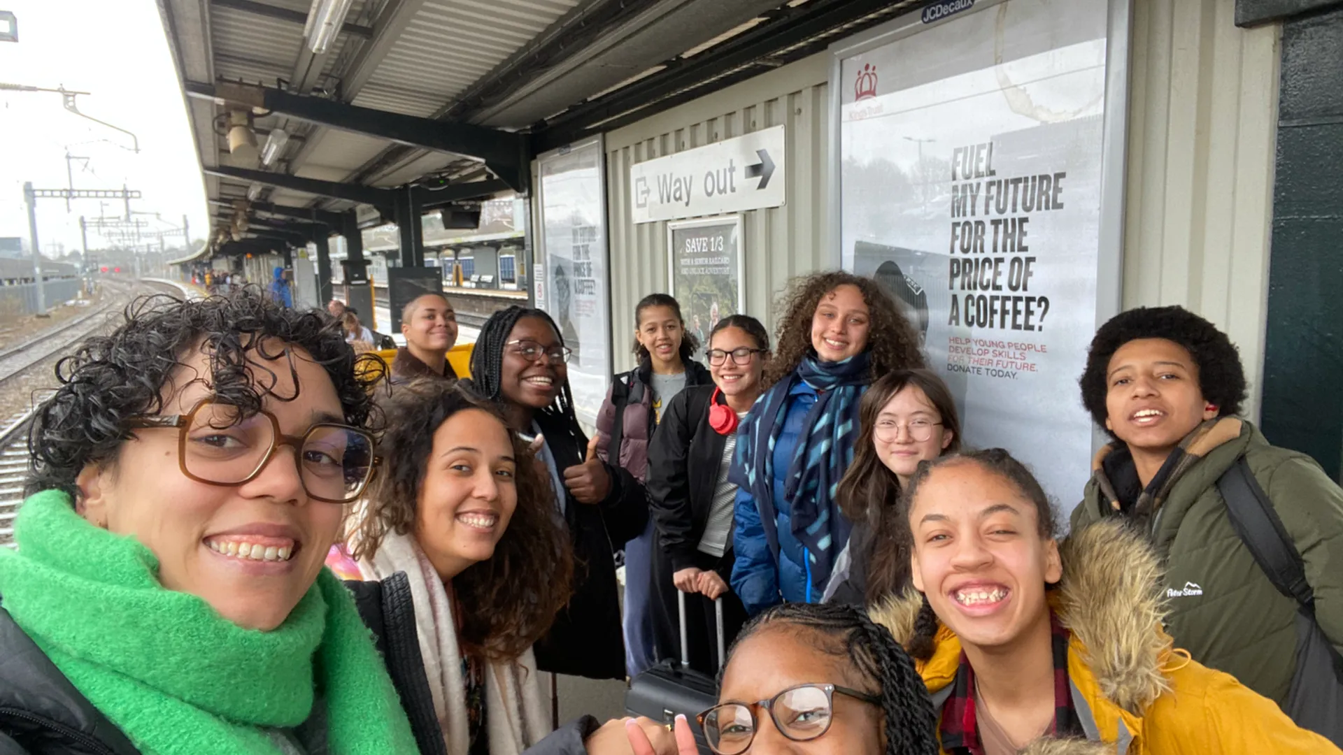A group of teenagers huddle close for a photograph. They're at a train station platform and smile alongside two women