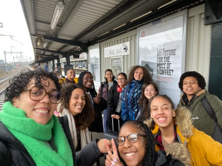 A group of teenagers huddle close for a photograph. They're at a train station platform and smile alongside two women