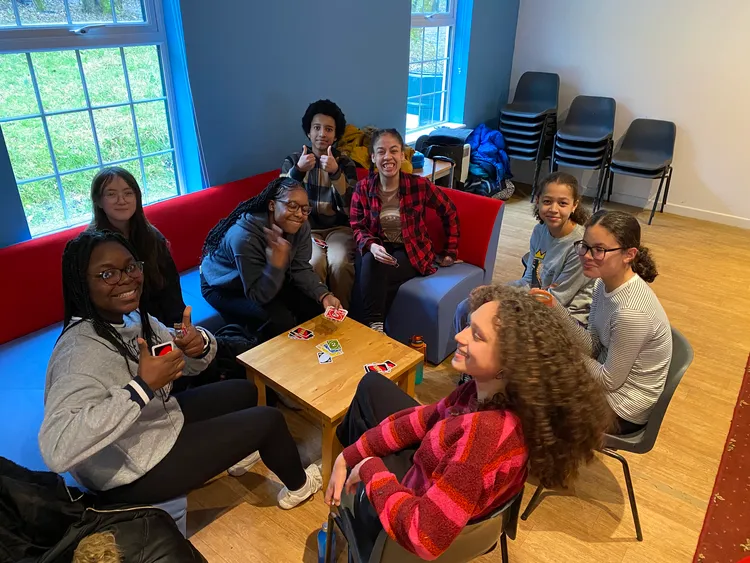 8 teens gather around a table, they smiles and show thumbs up towards a camera