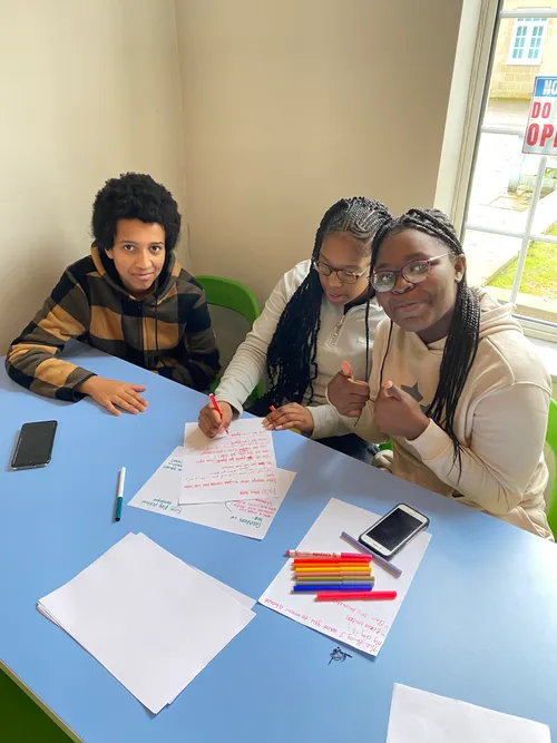 A boy and two girls in their mid-teens sit at a table with pens and paper, they're writing and smiling 