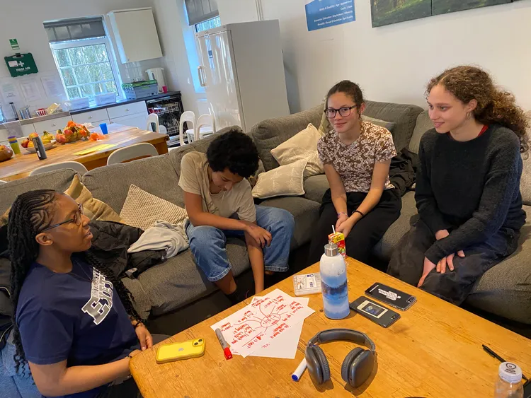 Four young people sit on a sofa, there's a sheet of paper and pens in front of them and they're talking to one another