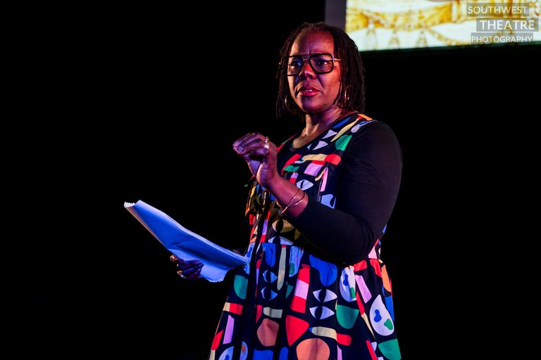A Black woman in a colourful dress holds a script and addresses the audience