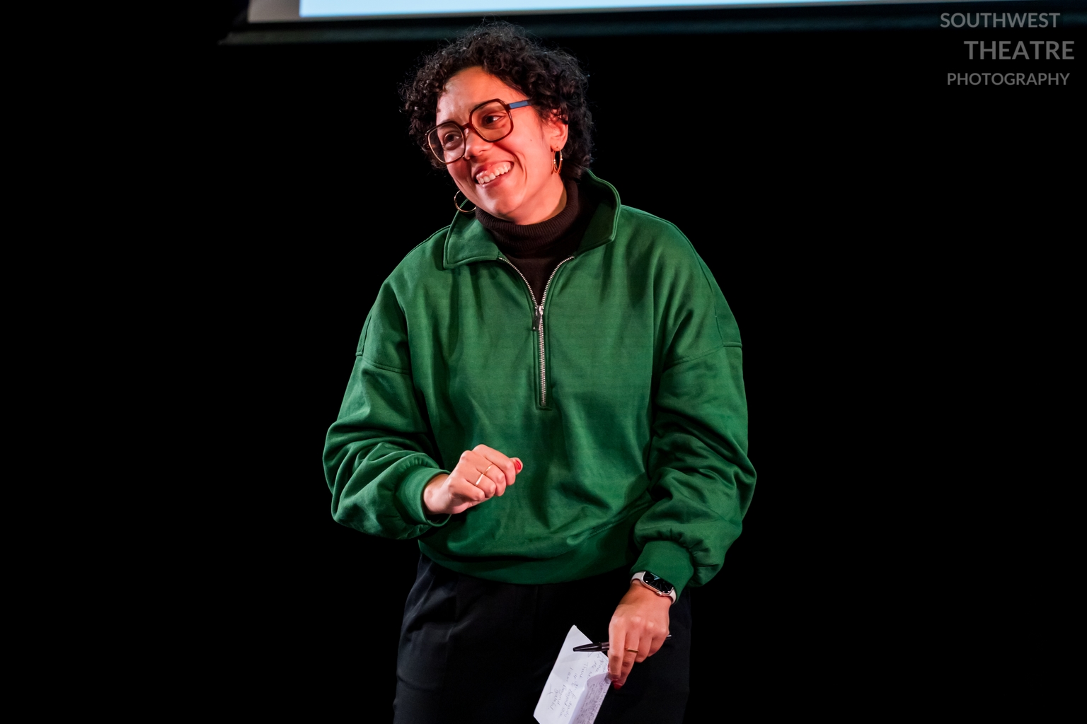A woman in a green jumper with short curly hair and glasses smiles at the audience