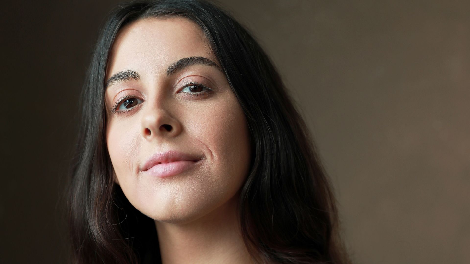 A woman with long brown hair smiles at the camera, she is wearing a brown top with a long silver necklace