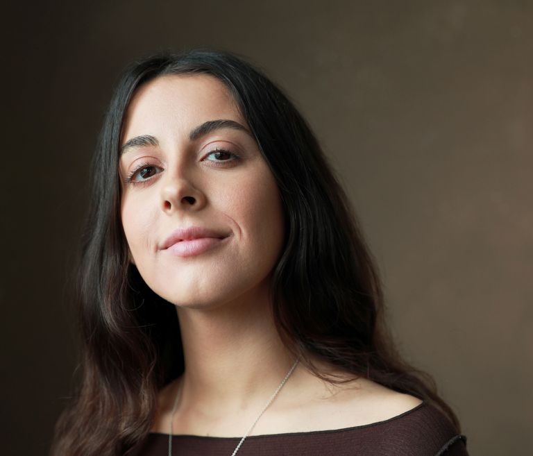A woman with long brown hair smiles at the camera, she is wearing a brown top with a long silver necklace