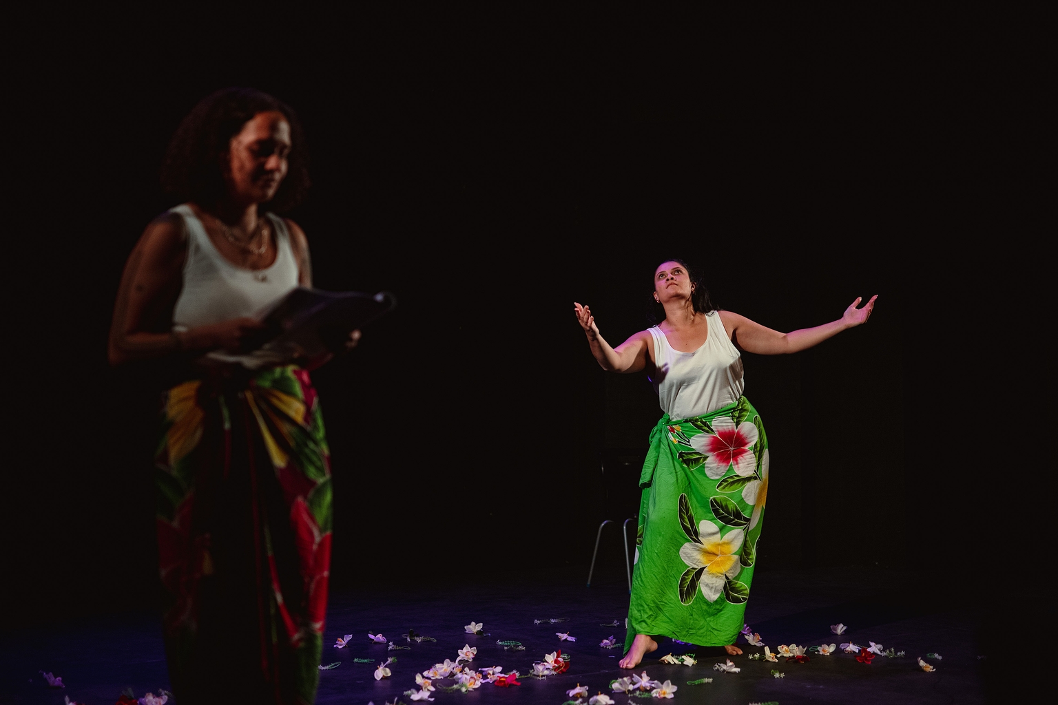 Two performers are on stage, surrounded my petals, wearing white t-shirts and flower print pareo's around their waists. One person is holding a script, the other is dancing in the back with her hands and head up towards the sky 
