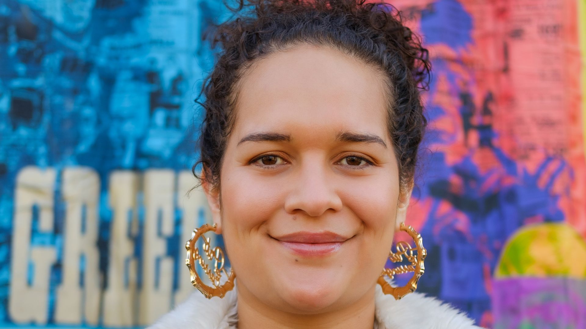 A woman with light brown curly hair smiles at the camera, she is wearing gold hoops and a green coat