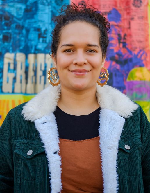 A woman with light brown curly hair smiles at the camera, she is wearing gold hoops and a green coat