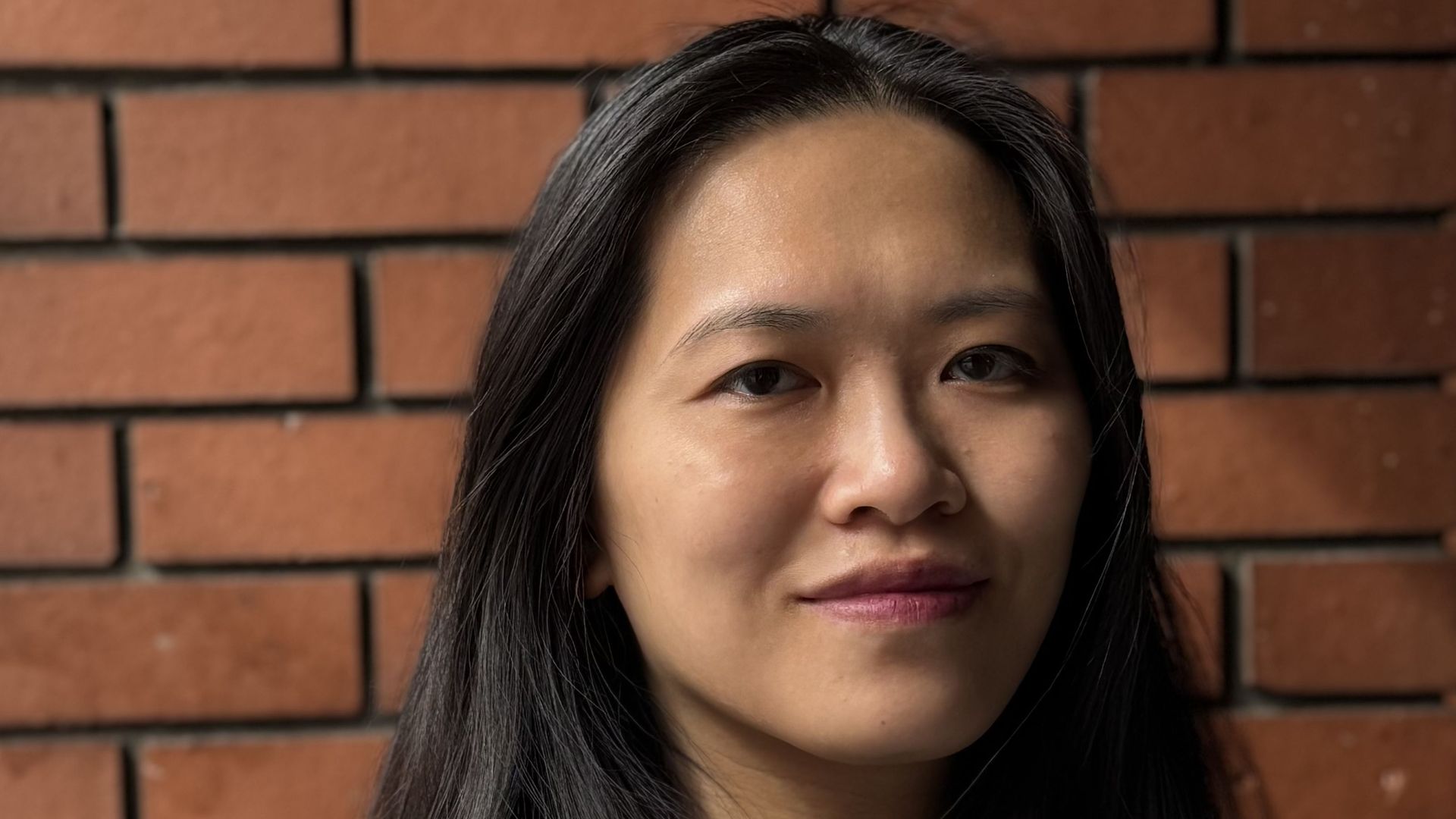 A woman with long brown hair stands against a brick wall gently smiling into the camera