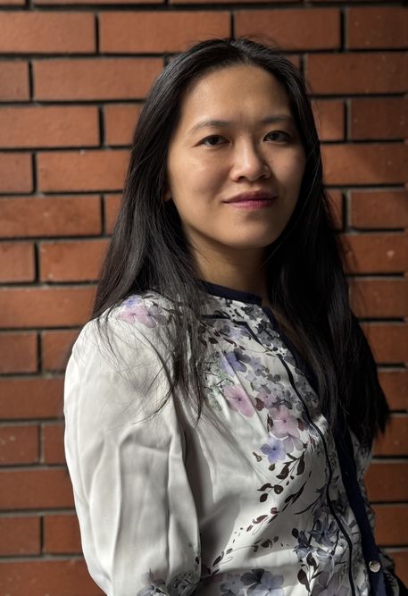 A woman with long brown hair stands against a brick wall gently smiling into the camera