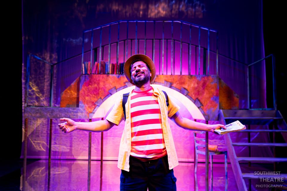 Person in a striped shirt and hat performing on a brightly lit stage, holding a script with arms outstretched in front of a colourful set.