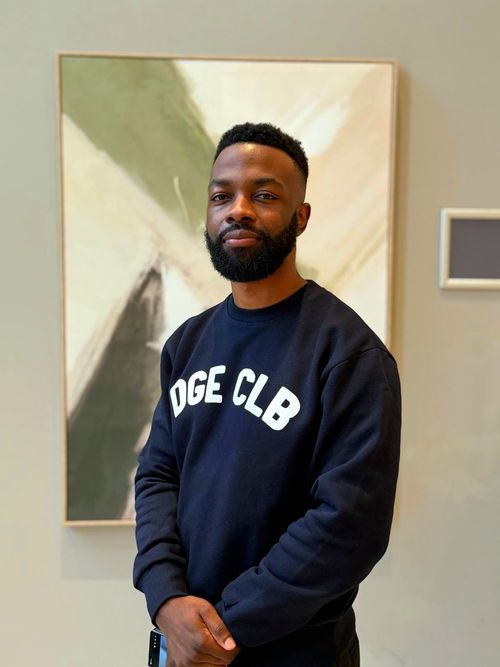 Photo of Associate Videographer Quason Matthews, standing in front of a framed piece of abstract art with their hands folded together in front of them