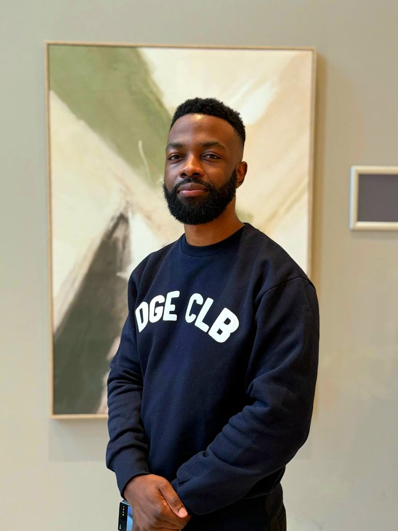 Photo of Associate Videographer Quason Matthews, standing in front of a framed piece of abstract art with their hands folded together in front of them