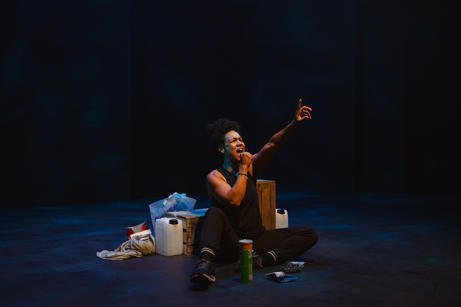 A performer sits on the floor of a stage, she is pretending to sing into a microphone and has one hand up in the air 