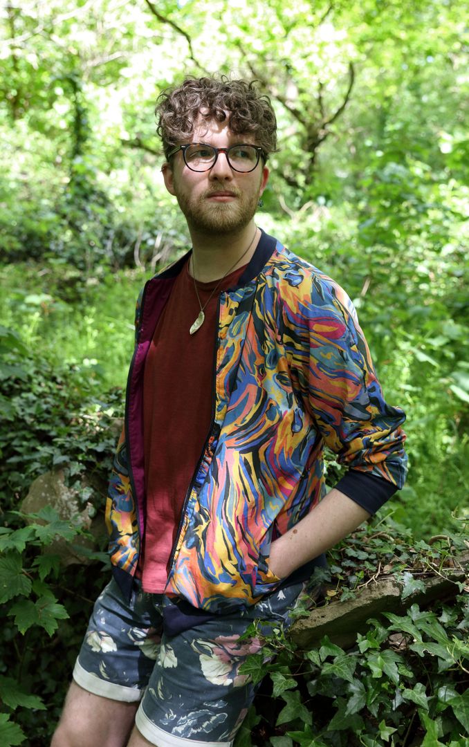 A man in a colour jacket is stood in a forest gazing into the distance