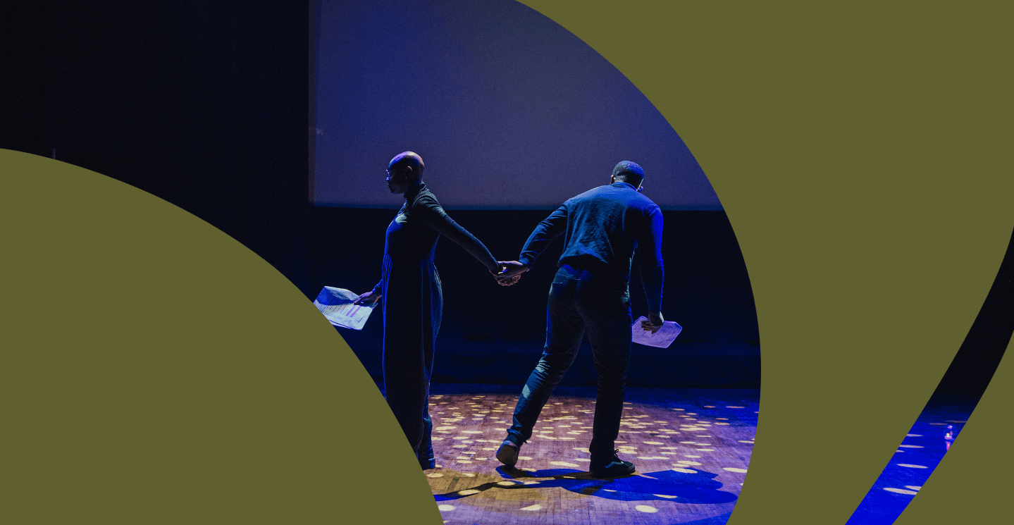 A man wearing a summer hat stands on a stage smiling with his eyes closed and his arms out either side holding a book