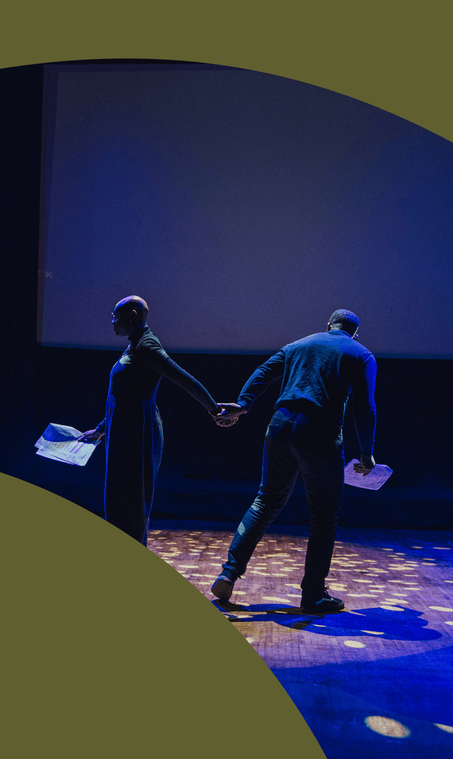 A man wearing a summer hat stands on a stage smiling with his eyes closed and his arms out either side holding a book