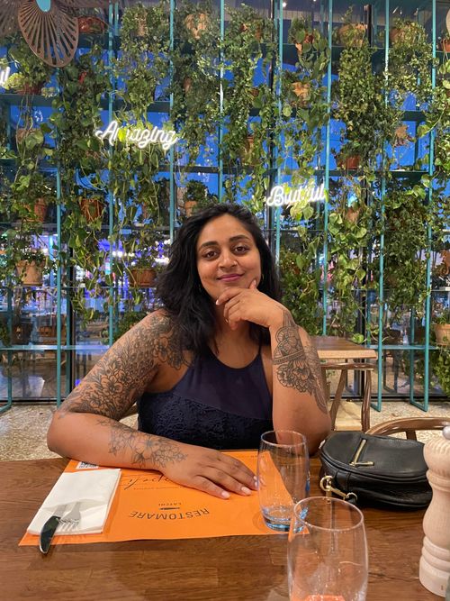 A woman with thick black hair and tattoos on her arms is smiling at a table in a restaurant, he left hand is leaning on her chin 