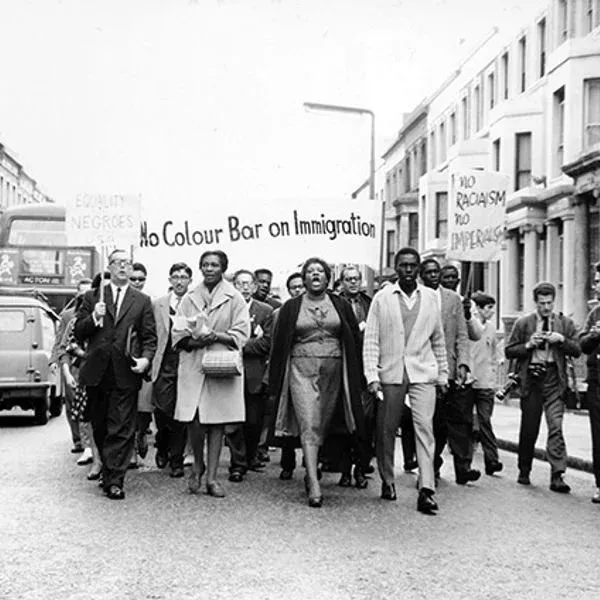 A black and white image showing a group of people of majority Black descent walking on the streets holding a banners reading 'No colour bar on immigration', 'No racism, no imperialism'