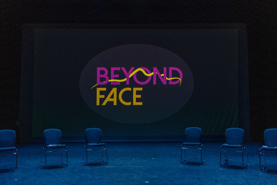 6 empty chairs are placed in a semi-circle on a stage, the old Beyond Face logo is projected onto the wall behind