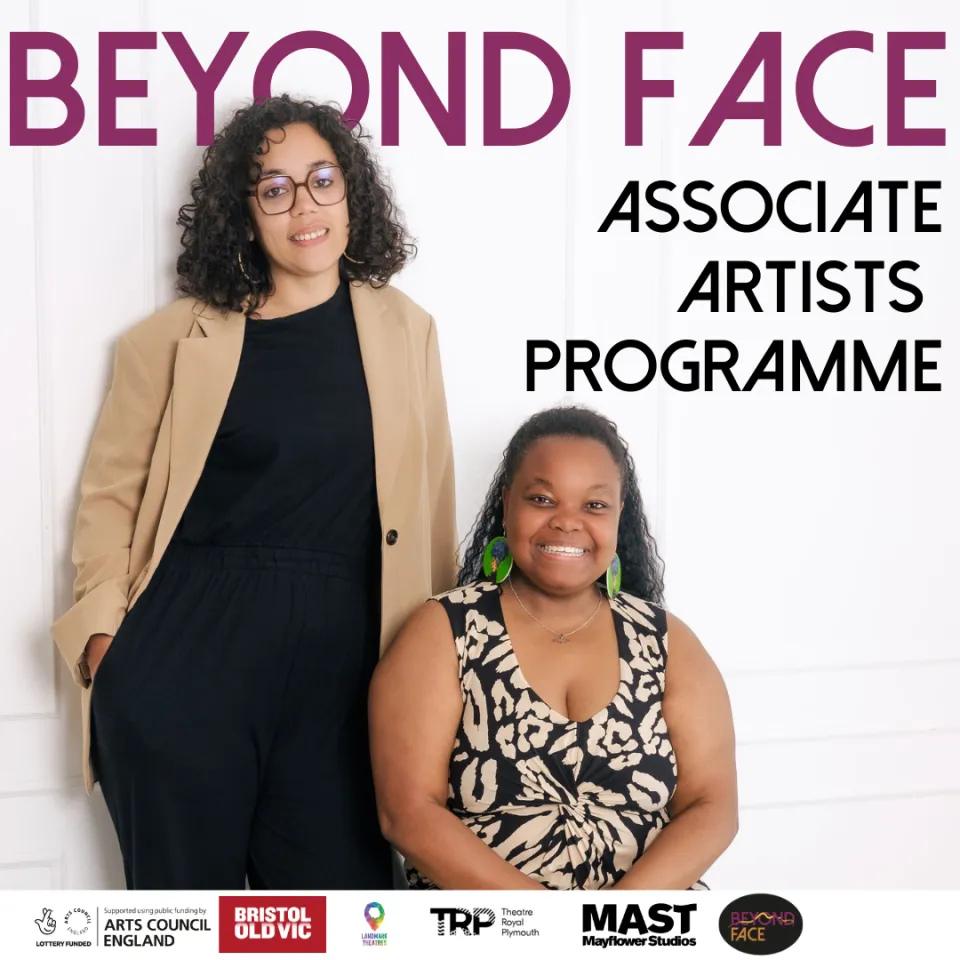 Two woman, one sitting, one standing smile towards a camera. The words Beyond Face Associate Artists Programme is written in the background