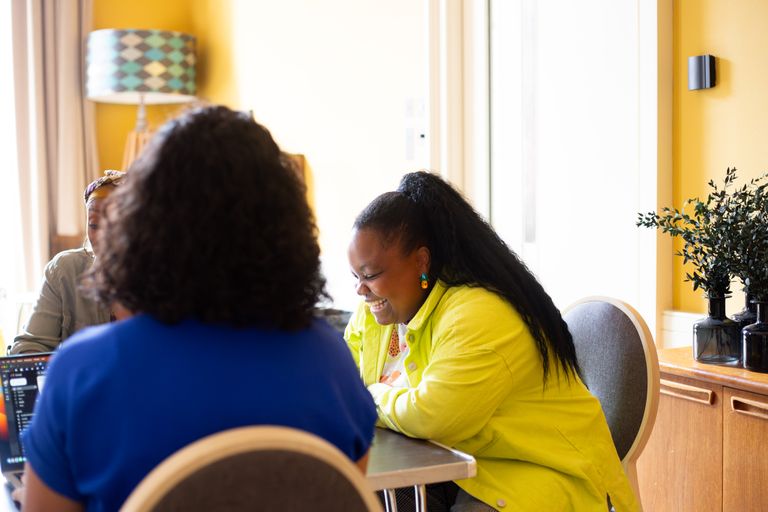 A woman with a long ponytail and yellow shirt sits at a desk laughing, there is another person in blue sat next to her
