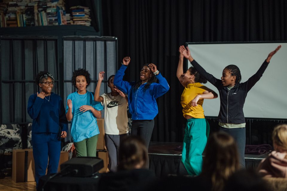 Group of young performers dancing and raising their arms on a small stage, with an audience watching in the foreground.