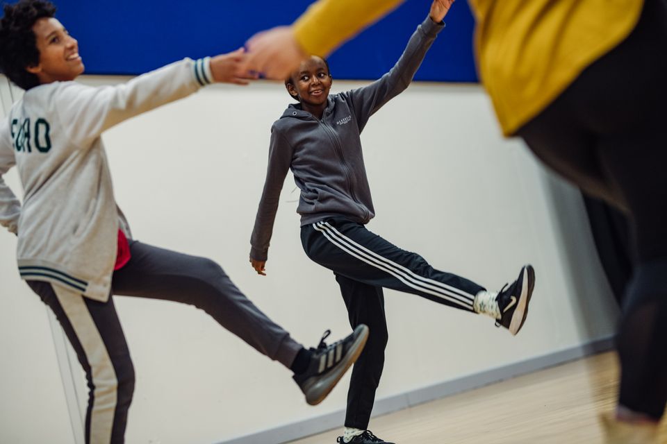 Young people taking part in a lively theatre workshop, raising arms and kicking legs as they move together in an exercise.