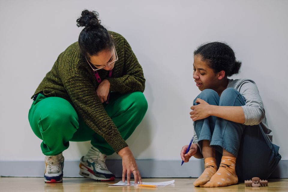 Photo of a young person sat on the floor with their knees up and a pen in their right hand, looking at a piece of paper on the floor with an adult who is squatted down next to them and pointing to something on the paper