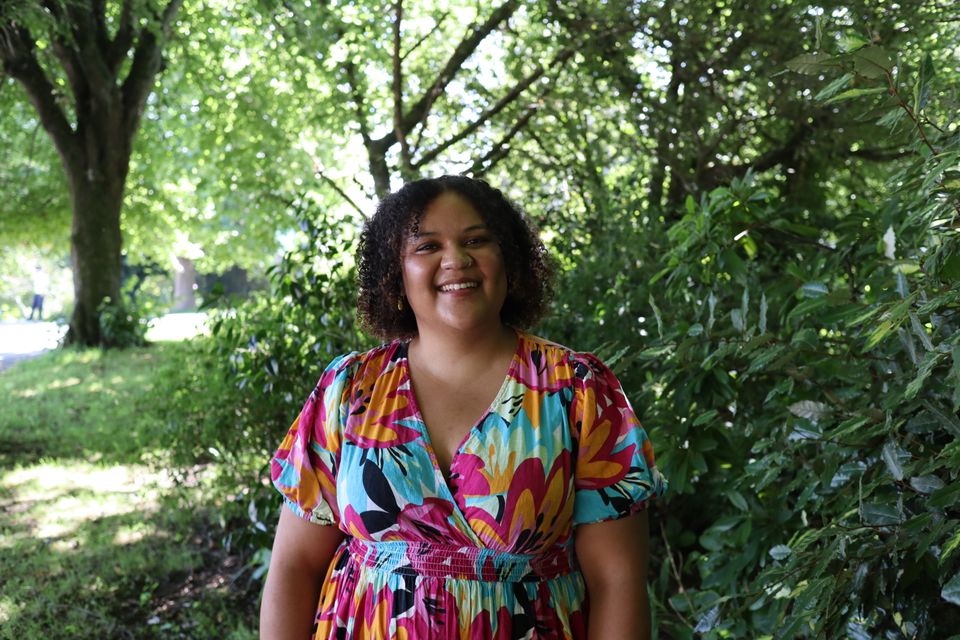Dulcie is stood near some trees smiling, they have curly brown hair and are wearing a colourful dress