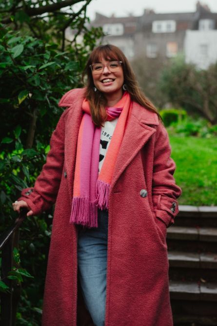 Hattie has a fringe and straight medium length hair, she is wearing a pink coat and matching scarf. She is smiling at the camera