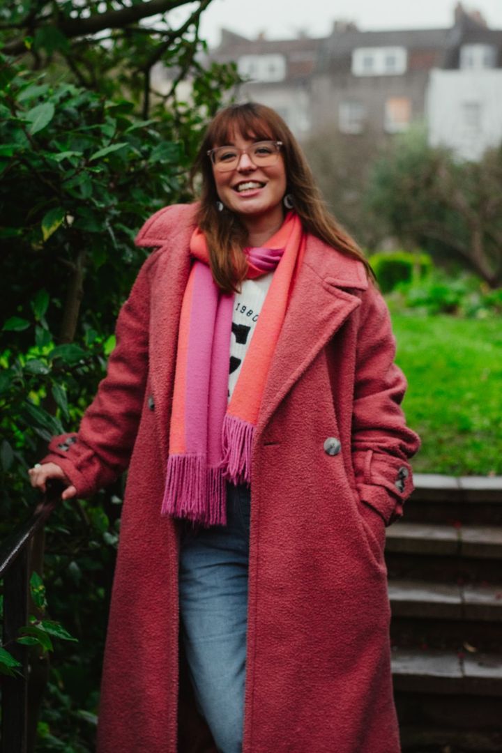 Hattie has a fringe and straight medium length hair, she is wearing a pink coat and matching scarf. She is smiling at the camera
