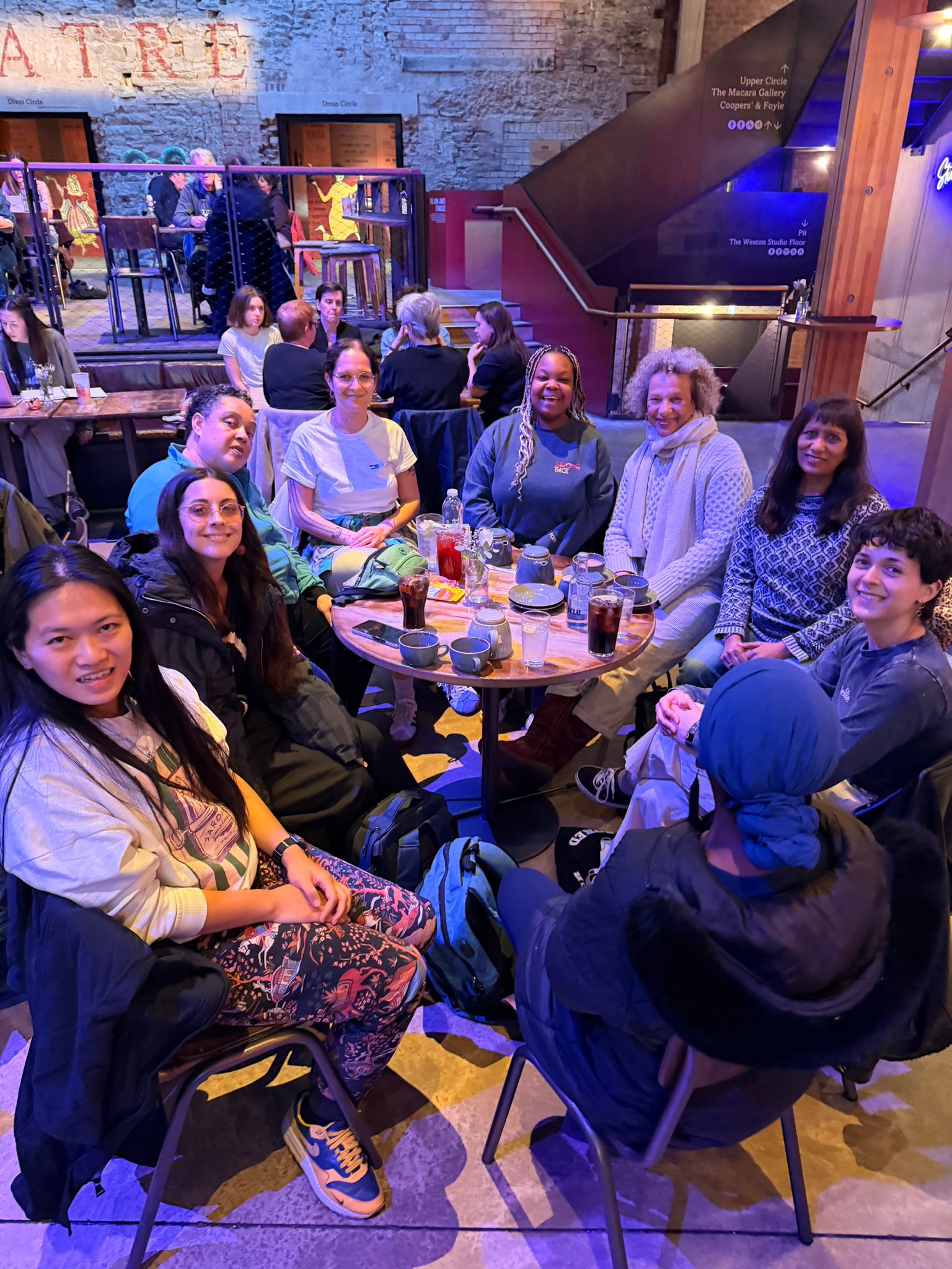 A group of nine people are sat around a table having drinks, they're smiling directly at the camera.