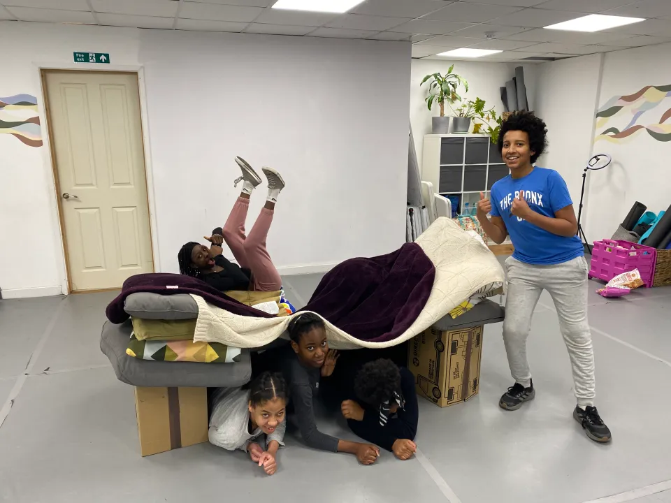 5 young people who have create a fort out of chairs pose for a photo underneath and ontop of the fort