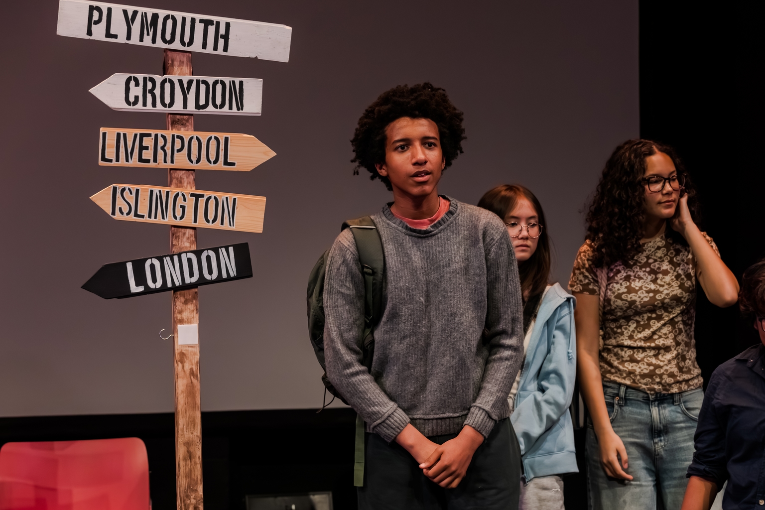 A young man stands on stage performing, behind him is a sign pointing in different directions to Plymouth, Croydon, Liverpool, Islington and London