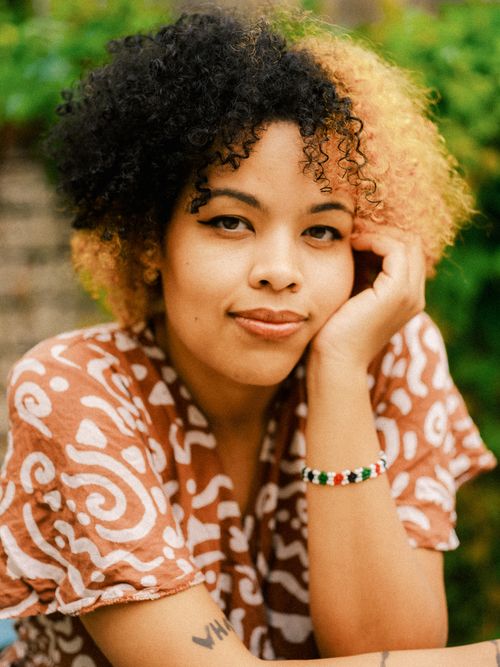 Malaika rests with one hand on her face, she has afro textured hair, half of it is dyed blonde