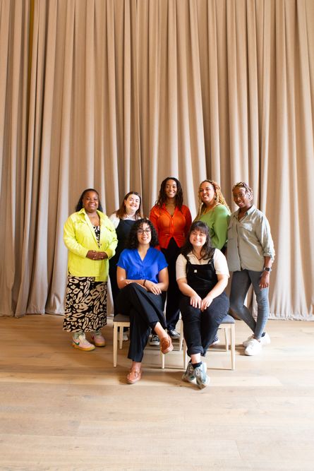 For a company group photo, 2 women are seated on chairs smiling and 5 more women stand behind them smiling towards the camera